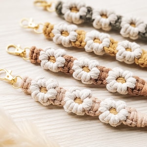 Könnte beinhalten: Nahaufnahme mehrerer Makramee-Blumen-Armbänder in verschiedenen Farben, darunter Beige, Braun und Senfgelb. Jedes Armband hat einen goldfarbenen Verschluss.