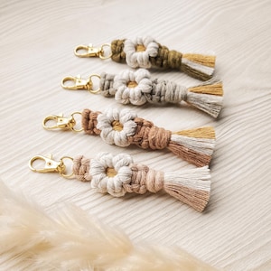 Puede incluir: Cuatro llaveros de macramé hechos a mano con cierres dorados. Cada llavero presenta un diseño floral y una borla en diferentes tonos de beige, marrón y verde. Los llaveros están dispuestos sobre una superficie de madera clara.