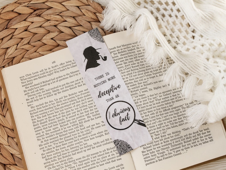 May include: A white bookmark featuring a black silhouette of a detective holding a pipe. The bookmark includes the text "There is nothing more deceptive than an obvious fact" and a magnifying glass with a fingerprint.