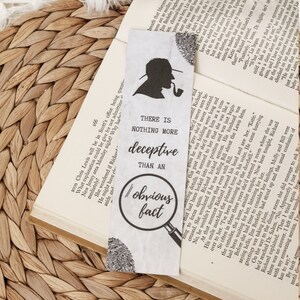 May include: A white bookmark featuring a black silhouette of a person wearing a hat and smoking a pipe. The bookmark has the text "There is nothing more deceptive than an obvious fact."