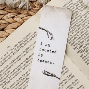 Puede incluir: Un marcap&aacute;ginas blanco con una ilustraci&oacute;n en blanco y negro de manos esquel&eacute;ticas. El marcap&aacute;ginas tiene el texto "I am haunted by humans." impreso en &eacute;l.