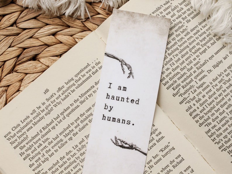 Puede incluir: Un marcap&aacute;ginas blanco con una ilustraci&oacute;n en blanco y negro de manos esquel&eacute;ticas. El marcap&aacute;ginas tiene el texto "I am haunted by humans." impreso en &eacute;l.