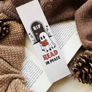 May include: A white bookmark with a Halloween theme. It features a tombstone with "RIP" and a ghost reading a book. The text "READ IN PEACE" is below the ghost. The bookmark is set on an open book.