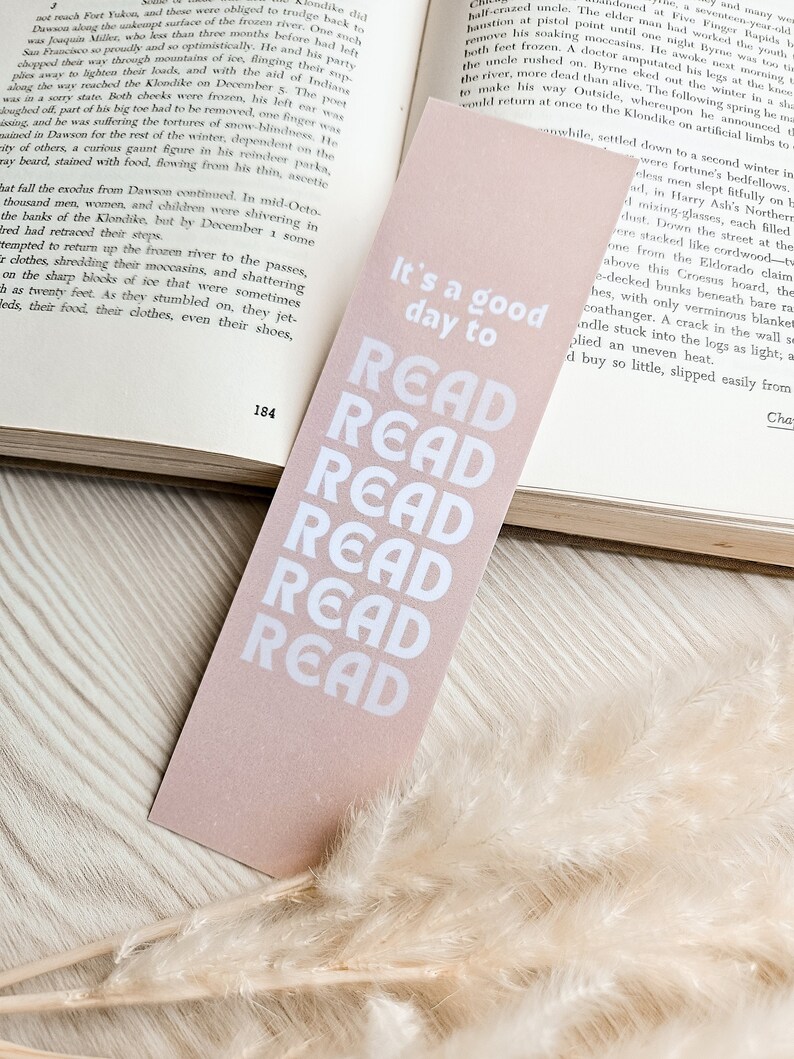 Pu&ograve; includere: Un segnalibro rettangolare rosa chiaro con la scritta bianca "It's a good day to READ" impilata verticalmente. Il segnalibro &egrave; posto su un libro aperto.