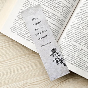 May include: A rectangular bookmark with the quote "Hell is empty and all the devils are here" by Shakespeare. The bookmark features a black and white illustration of a rose and a skeletal hand. The bookmark is placed in an open book.