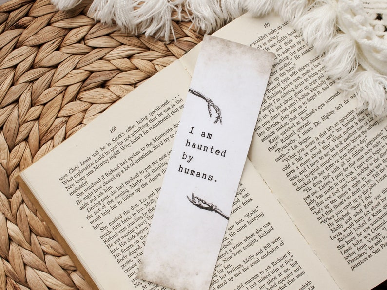 Puede incluir: Un marcap&aacute;ginas blanco con un dise&ntilde;o de esqueleto negro y el texto "Estoy obsesionado por los humanos". El marcap&aacute;ginas est&aacute; sobre un libro con p&aacute;ginas marrones.