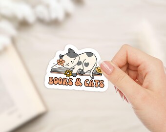 Könnte beinhalten: Ein Aufkleber mit einer Cartoon-Katze mit grau-weißen Flecken, die auf einem offenen Buch liegt. Der Aufkleber trägt den orangefarbenen Schriftzug "BOOKS & CATS" mit Blumen.