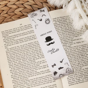 Puede incluir: Un marcap&aacute;ginas en blanco y negro con ilustraciones tem&aacute;ticas de detectives, incluyendo un sombrero, un bigote y una lupa. El marcap&aacute;ginas tiene el texto "Hercule Poirot" y "Agatha Christie". Est&aacute; colocado en un libro abierto.