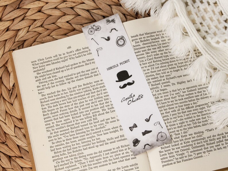 Puede incluir: Un marcap&aacute;ginas en blanco y negro con ilustraciones tem&aacute;ticas de detectives, incluyendo un sombrero, un bigote y una lupa. El marcap&aacute;ginas tiene el texto "Hercule Poirot" y "Agatha Christie". Est&aacute; colocado en un libro abierto.