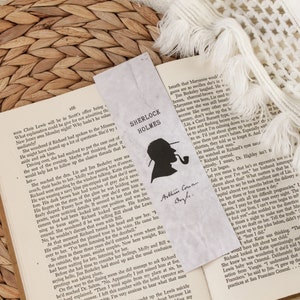 May include: A close-up of an open book with a bookmark featuring a silhouette of Sherlock Holmes. The bookmark has the text "Sherlock Holmes" and "Arthur Conan Doyle" written on it. The book is open to a page with text about a detective story.