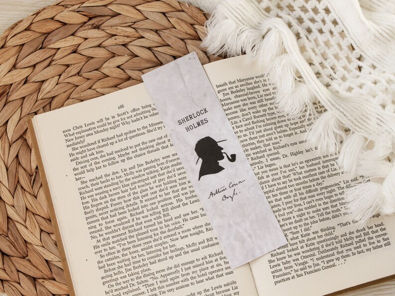 May include: A close-up of an open book with a bookmark featuring a silhouette of Sherlock Holmes. The bookmark has the text "Sherlock Holmes" and "Arthur Conan Doyle" written on it. The book is open to a page with text about a detective story.