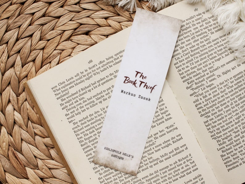 Puede incluir: Un marcap&aacute;ginas marr&oacute;n y blanco con el t&iacute;tulo "El ladr&oacute;n de libros" de Markus Zusak. El marcap&aacute;ginas est&aacute; hecho de papel y tiene un aspecto vintage.