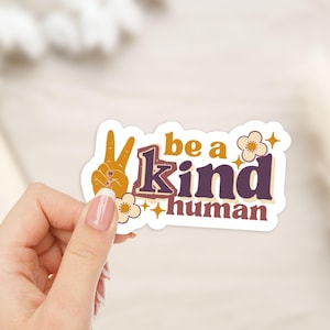 Può includere: Un adesivo con la scritta "be a kind human" in un font retrò. L'adesivo presenta un gesto della mano del segno della pace in arancione, con fiori e stelle. Lo sfondo è di un colore neutro.