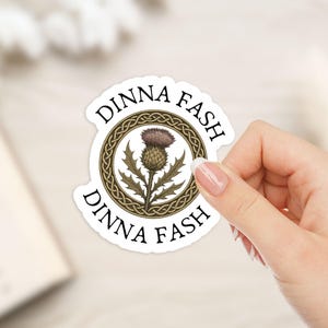 May include: A white sticker with a thistle design and the words "DINNA FASH" in a circle. The thistle is brown and purple, set within a gold Celtic knot. The sticker is held by a hand.