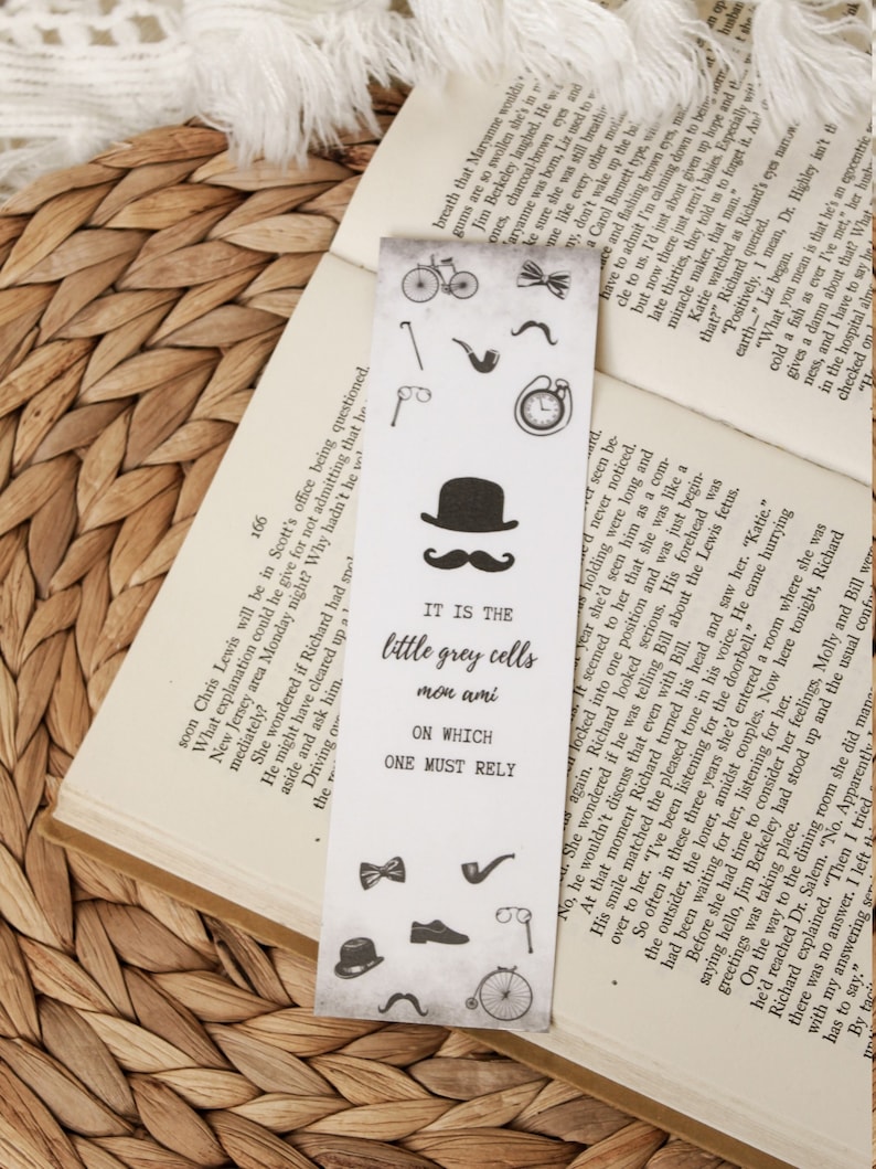 Puede incluir: Un marcap&aacute;ginas de estilo vintage con un fondo gris claro, con varias ilustraciones negras de una bicicleta, una pipa, un sombrero, un bigote y el texto "IT IS THE little grey cells mon ami ON WHICH ONE MUST RELY".