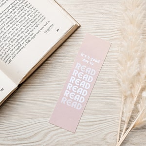 Pu&ograve; includere: Un segnalibro rosa pallido con testo bianco che recita "It's a good day to READ" impilato verticalmente. Il segnalibro &egrave; posizionato vicino a un libro aperto e a erba della pampa essiccata.