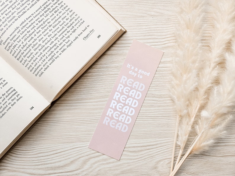 Pu&ograve; includere: Un segnalibro rosa pallido con testo bianco che recita "It's a good day to READ" impilato verticalmente. Il segnalibro &egrave; posizionato vicino a un libro aperto e a erba della pampa essiccata.