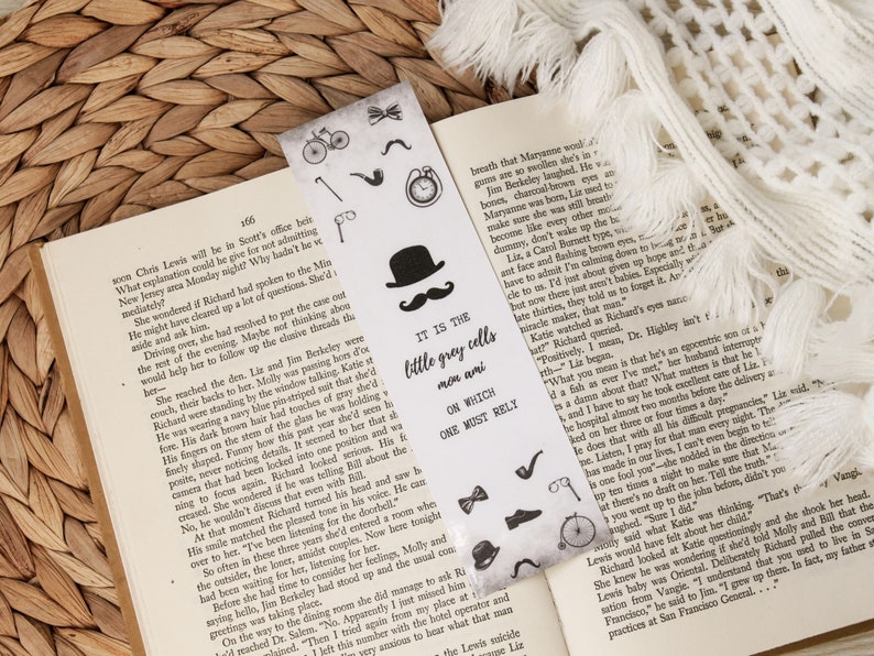 Puede incluir: Marcap&aacute;ginas de dise&ntilde;o vintage con sombrero, bigote, pipa y otros iconos de caballero. El marcap&aacute;ginas tiene el texto "IT IS THE little grey cells men ami ON WHICH ONE MUST RELY" y est&aacute; colocado en un libro abierto.