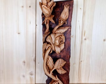 May include: A carved wooden wall decoration depicting roses and leaves. The roses are at different stages of opening, with detailed carving. The background is a dark brown, rectangular piece of wood, and the roses are a lighter, natural wood colour.