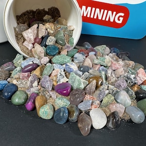 Puede incluir: Una colección de piedras preciosas pulidas de colores que se derraman de un recipiente blanco sobre una superficie oscura. Las piedras preciosas varían en tamaño, forma y color, incluyendo tonos de azul, verde, púrpura, rosa y marrón. La palabra "MINING" es visible.