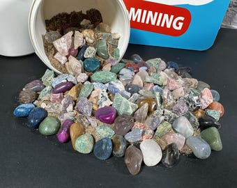 Puede incluir: Una colección de piedras preciosas pulidas de colores que se derraman de un recipiente blanco sobre una superficie oscura. Las piedras preciosas varían en tamaño, forma y color, incluyendo tonos de azul, verde, púrpura, rosa y marrón. La palabra "MINING" es visible.