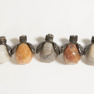 Może przedstawiać: Pięć małych figurek żółwi wyrzeźbionych z różnych kolorowych kamieni. Żółwie są skierowane w tym samym kierunku i mają szary pancerz z białymi oznaczeniami. Kamienie są białe, pomarańczowe, szare, czerwone i różowe.