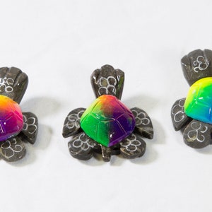 May include: Three small wooden turtle figurines with rainbow colored shells. The turtles are carved from wood and have a natural brown color. The shells are painted with a rainbow of colors, including red, orange, yellow, green, blue, indigo, and violet.