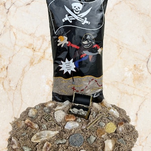 Puede incluir: Una bolsa de plástico negra con un diseño de pirata y el texto "Discover Buried Treasures Inside". La bolsa contiene un montón de arena marrón con un pequeño cofre del tesoro dorado, monedas de oro y una llave.