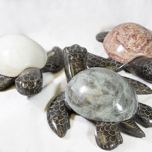 May include: Three stone turtle sculptures with detailed flipper and shell designs. One turtle has a white shell, another a reddish-brown shell, and the third a gray shell. The turtles are on a white background.