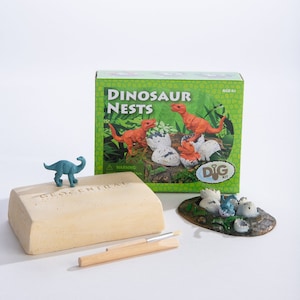 Pode incluir: Kit de escavação de ninhos de dinossauros com uma figura de dinossauro verde, um pincel marrom, um bloco de argila bege e uma caixa com fundo verde e o texto "Dinosaur Nests" e "Dig Kit".