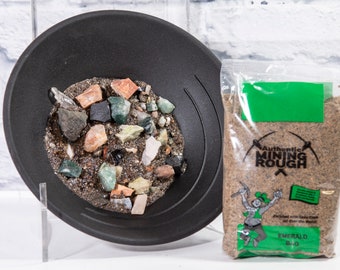 May include: A black plastic gold pan filled with small rocks and minerals. A clear plastic bag with a green label reads "Authentic Mining Rough" and "Emerald Dig".