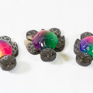 May include: Three small, colorful, wooden turtle figurines. Each turtle is painted with a rainbow gradient, with the colors blending from yellow to green to purple. The turtles are carved from wood and have a detailed shell and head.