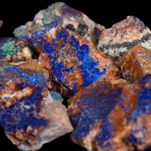 May include: A close-up of a group of rough, raw, blue azurite crystals with brown and green accents. The crystals are clustered together and have a natural, earthy appearance.