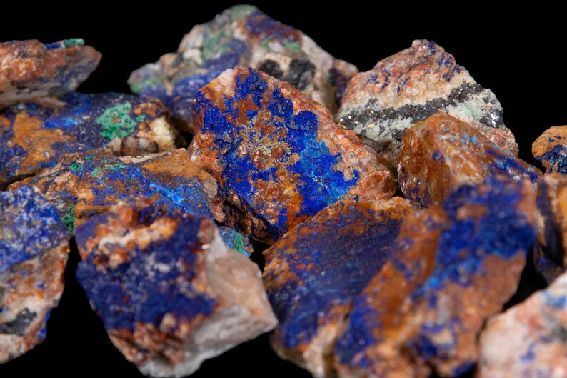 May include: A close-up of a group of rough, raw, blue azurite crystals with brown and green accents. The crystals are clustered together and have a natural, earthy appearance.