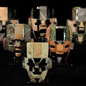 May include: Six colorful stone masks with geometric designs. The masks are made of different types of stone, including green, black, and brown. They are all carved in a similar style, with large eyes and a wide mouth.
