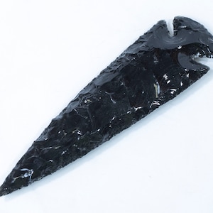 May include: A black obsidian arrowhead with a pointed tip and a notch at the top. The arrowhead is made of a dark, glassy material and has a smooth, polished surface.