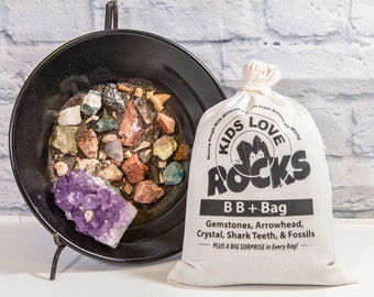 May include: A white canvas bag with the text "Kids Love Rocks" and the words "BB + Bag" printed on it. The bag contains a variety of gemstones, arrowheads, crystals, shark teeth, and fossils. The bag is sitting on a black pan with a purple amethyst crystal.
