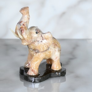 May include: A small, hand-carved elephant figurine made of light brown and white marble. The elephant is standing on a black base and has its trunk raised in the air.