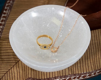 Könnte beinhalten: Eine weiße Selenit-Schale mit einem goldenen Ring und einer goldenen Kettenkette mit einem kleinen Kristall-Anhänger. Die Schale steht auf einem braunen und blauen gemusterten Stoff.