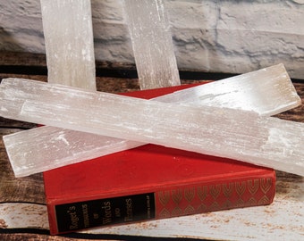 May include: Three white selenite sticks, also known as selenite wands, are stacked on top of a red book with gold lettering. The book is titled "Roget's Thesaurus of Words and Phrases".