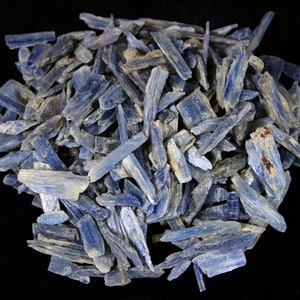 May include: A pile of blue kyanite crystal chips. The chips are long and thin, with a slightly rough texture. They are a light blue color, with some darker blue streaks.