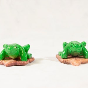 May include: Two small, green frog figurines with speckled details, resting on reddish-brown leaf-shaped bases. The frogs are positioned side-by-side against a plain white background, showcasing their vibrant color and design.