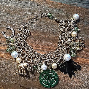 May include: A gold chain bracelet with multiple charms, including a horseshoe, elephants, a four-leaf clover, and a green jade pendant with Chinese characters. The bracelet also features white pearls and green glass beads.