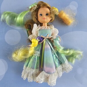 May include: A doll with brown hair featuring green and yellow highlights, styled in pigtails with plastic accessories. The doll wears a dress with a pastel tie-dye pattern, white lace trim, sheer puff sleeves, and a thin ribbon at the waist.