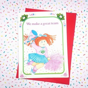 May include: A white card with a green border features a cartoon illustration of a red-haired girl wearing a blue and white cheerleader outfit. She is holding a megaphone and a pink pom-pom. The text "We make a great team" is printed in pink on the card.