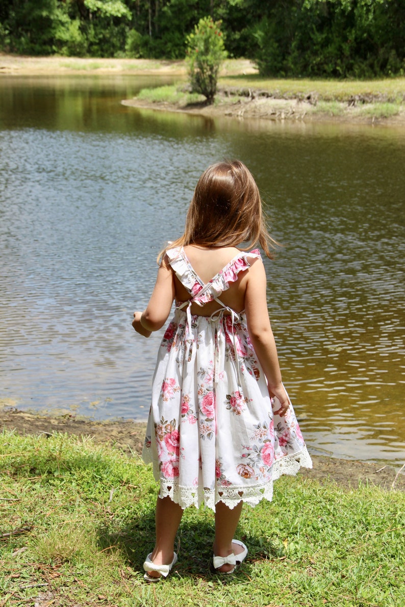 May include: A young girl wearing a white floral dress with pink flowers and lace trim stands by a pond. She is looking out at the water.