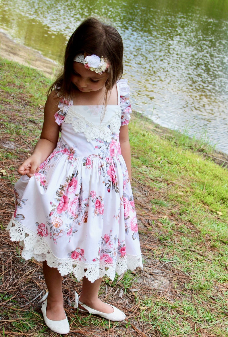 May include: A young girl wearing a white dress with pink floral print and lace trim. She is wearing a white headband with a flower accent. The dress has a square neckline and a gathered skirt. The girl is standing in a grassy area near a body of water.