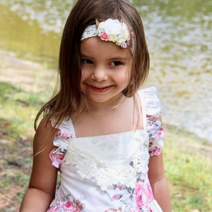 May include: A young girl wearing a white floral dress with a lace trim and a white headband with pink and yellow flowers. She is smiling and looking at the camera.