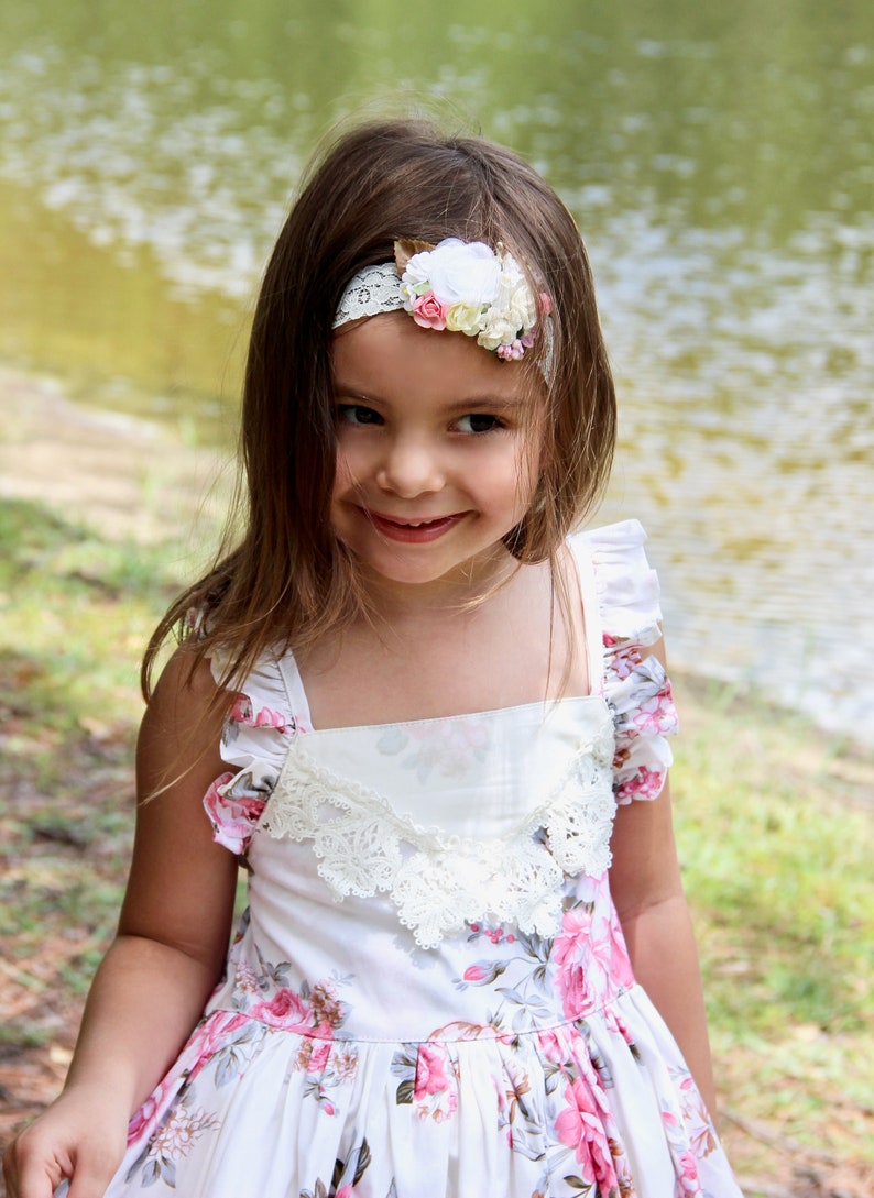 May include: A young girl wearing a white floral dress with a lace trim and a white headband with pink and yellow flowers. She is smiling and looking at the camera.