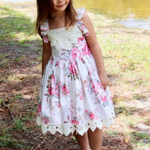 May include: A young girl wearing a white dress with pink and gray floral print and a white lace trim. She is wearing a white headband with a floral design and white shoes. The girl is standing in a grassy area with pine needles on the ground.